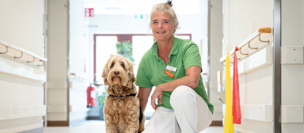 En medarbetare i vården med grön tröja knäböjer bredvid en brun hund.